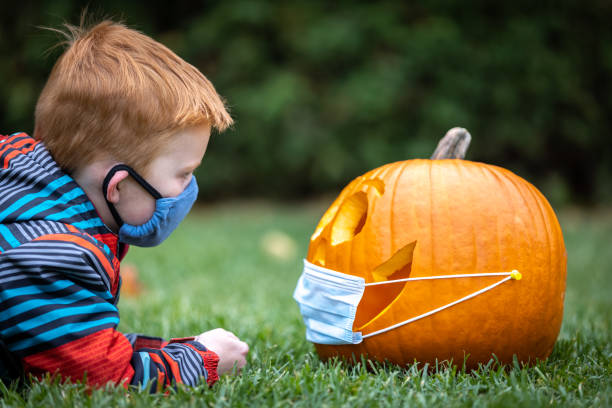 istock Cute redhead boy looking a Jack O' Lantern while lying down on grass and wearing wearing protective mask 1280982525