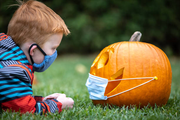 istock Cute redhead boy looking a Jack O' Lantern while lying down on grass and wearing wearing protective mask 1280982558