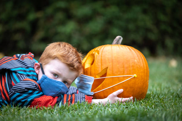 istock Cute redhead boy looking a Jack O' Lantern while lying down on grass and wearing wearing protective mask 1280989219