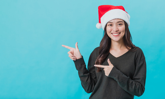 Happy young Asian woman wearing Santa Claus and hand pointing with happy smiling face isolated on blue background in studio shot.Christmas and happy new year celebration concept.