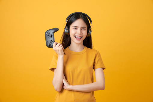 Cheerful beautiful Asian woman in casual yellow t-shirt and playing video games using joysticks with headphones on orange background.