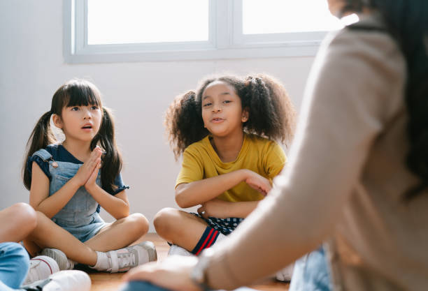 studenti internazionali delle elementari seduti sul pavimento in cerchio intorno all'insegnante e ascoltando una storia nella loro classe. l'insegnante sta leggendo la storia di un libro. istruzione, scuola,concetto di apprendimento - floor sitting foto e immagini stock