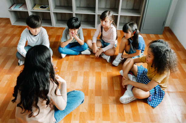 初級留学生グループは、先生の周りに円を描いて足を組んで座り、話を聞きます。学校の教室での子供と教師のディスカッショングループ。教育、学習コンセプト - eight legged ストックフォトと画像
