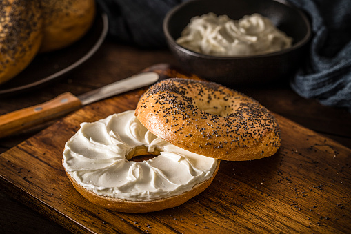 High angle view of an opened poppy seeds bagel spread with cream cheese. The bagel is on a rustic wooden table and it's surrounded by a black plate with two bagels and a bowl with cream cheese. Low key DSLR photo taken with Canon EOS 6D Mark II and Canon EF 100 mm f/ 2.8
