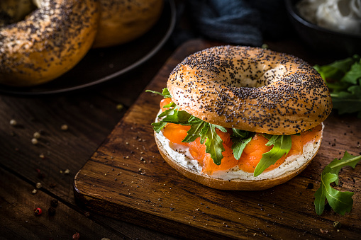 High angle view of a poppy seeds bagel spread with cheese cream and with smoked salmon and arugula topping on a rustic wooden table. The bagel is at the right side of the image and at the top left corner are two defocused bagels on a black plate. Low key DSLR photo taken with Canon EOS 6D Mark II and Canon EF 100 mm f/ 2.8