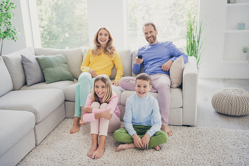 Full length photo of comfort family watch tv hold remote control two small kids, sit floor mom dad on couch in house indoors
