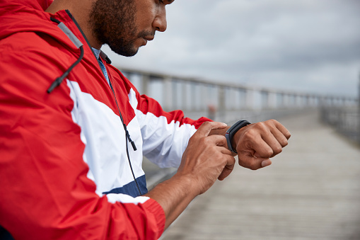 African-american young man with earphones wearing wind jacket tapping fitness tracker by river bridge