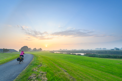 Cyclist riding on a levee during sunrise over the IJsseldelta near Kampen in Overijssel. The road on the levee between Kampen and Zwolle is winding into the distance where old farms are situated on the side of the dyke. Wide panoramic photo