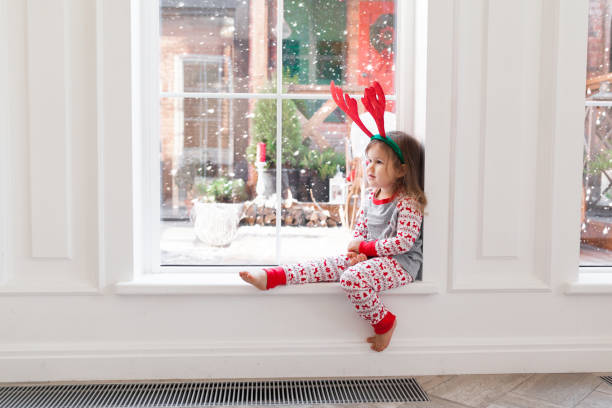 portret słodkiej dziewczyny w piżamie xmas i poroża reniferów siedzi przy oknie w zimie. świąteczny sezon weekendowy z rodziną w domu - christmas window magic house zdjęcia i obrazy z banku zdjęć