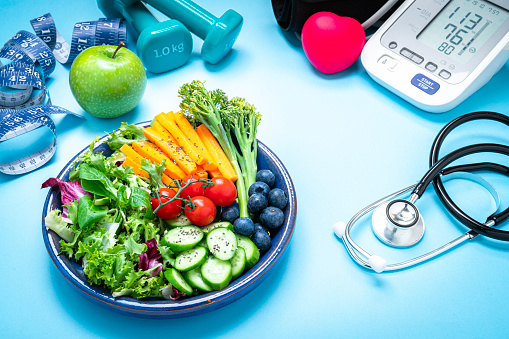 Healthy lifestyle concept: dumbbells, tape measure, salad plate, digital blood pressure monitor, doctor stethoscope and red heart shot on blue background. High resolution 42Mp studio digital capture taken with SONY A7rII and Zeiss Batis 40mm F2.0 CF lens