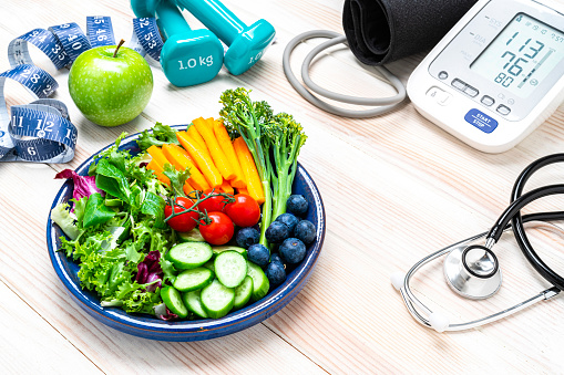 Healthy lifestyle concept: dumbbells, tape measure, salad plate, digital blood pressure monitor and doctor stethoscope shot on white table. High resolution 42Mp studio digital capture taken with SONY A7rII and Zeiss Batis 40mm F2.0 CF lens