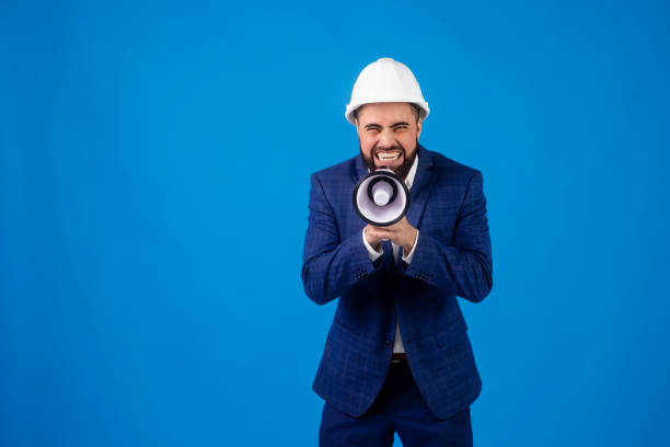 a bearded middle-aged construction worker in a suit and construction helmet speaks into a megaphone on a blue isolated background . a young architectural engineer shouts over a loudspeaker - home addition audio imagens e fotografias de stock