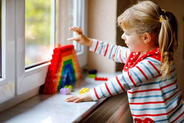 istock Cute little toddler girl by window create rainbow with colorful plastic blocks during pandemic coronavirus quarantine. Children made and paint rainbows around the world as sign. 1281074005