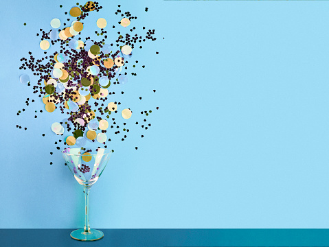 Close-up of martini glass splashing confetti. It is on colored background. Image represents festive celebration.