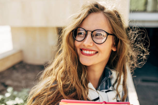 Closeup portrait of a smiling young student woman wearing transparent eyeglasses standing next to the college campus and carrying lots of books and folders on a sunny day. stock photo