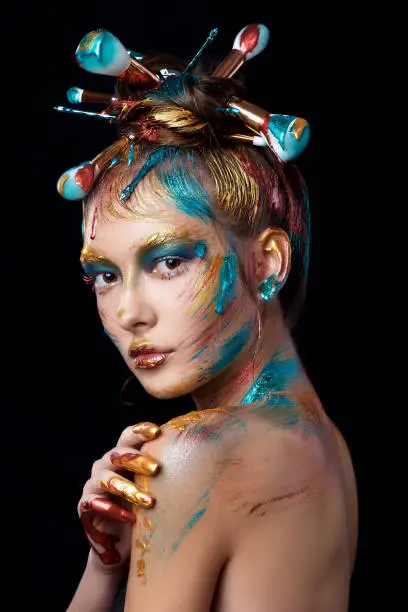 Close-up portrait of a young girl on a black background. Beautiful model with brushes for make-up in hair. Advertising space. Multicolor creative make-up. Color paint on the face and hair.