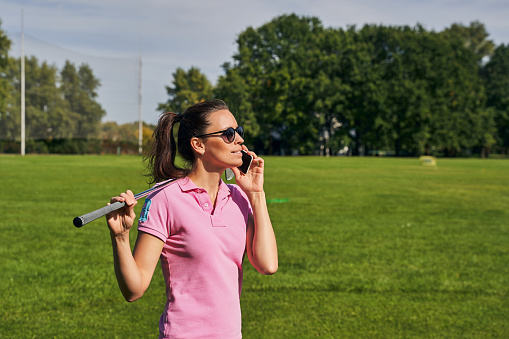 Beautiful dark-haired young Caucasian female golfer in round sunglasses calling on her cellular phone outdoors