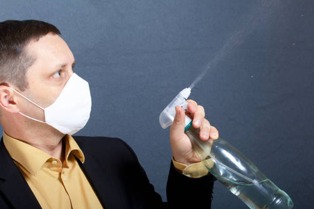 A man in a suit without a tie. Medical mask on the face. Demonstrates the operation of a spray bottle with an antiseptic. In front of him is a stand for a smartphone with a lamp for illumination. Video communication. un hombre con traje sin corbata. máscara médica en la cara. demuestra el funcionamiento de una botella de pulverización con un antiséptico. frente a él hay un soporte para un teléfono inteligente con una lámpara para la iluminación. comunicación p