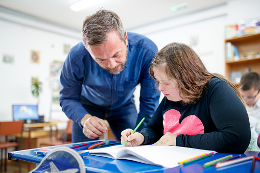 A girl with down syndrome is in a classroom with her teacher, she is drawing something in her notebook while being watched by her teacher