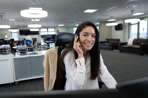 Portrait of mixed race businesswoman working in a modern office, sitting at desk talking using phone headset and smiling to camera. Social distancing in workplace during Coronavirus Covid 19 pandemic.