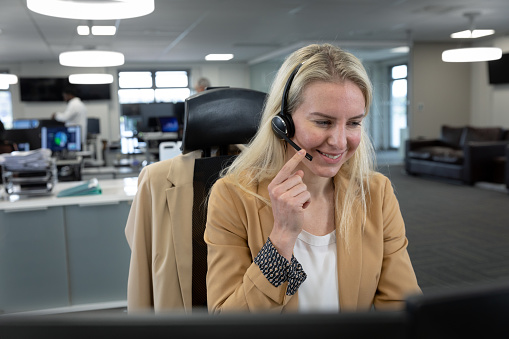 Caucasian businesswoman working in a modern office, sitting at desk talking using phone headset and smiling. Social distancing in workplace during Coronavirus Covid 19 pandemic.