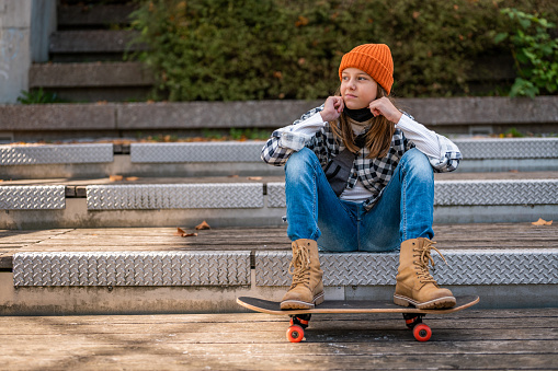 Portrait of a young cool girl with waist bag, wearing protective face mask under her chin while sitting on the skateboard outdoors on some staircase.