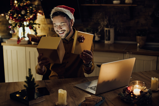 Young happy man opening Christmas present while having video call over a computer in the evening at home.
