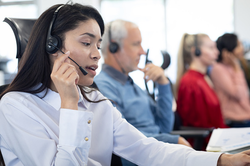 Mixed race businesswoman working in a modern office, sitting at desk talking using phone headset, with colleagues wearing headsets in the background. Business communications team at work.