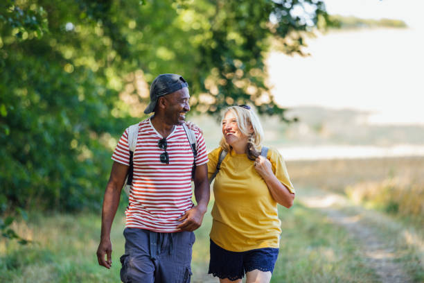 Time with Her! A multi-ethnic senior couple wearing casual clothing and backpacks walking down a footpath in a rural forest on a bright day in Alnwick. They are laughing together. habitat 67 stock pictures, royalty-free photos & images