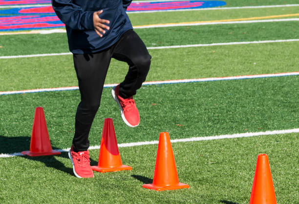 Athlete stepping over orange cones on a turf field A high school track and field sprinter performs running drills over orange cones on a green turf field. sports training drill stock pictures, royalty-free photos & images