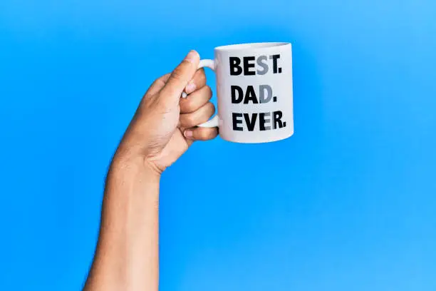 Hand of hispanic man holding best dad ever coffee cup over isolated blue background.