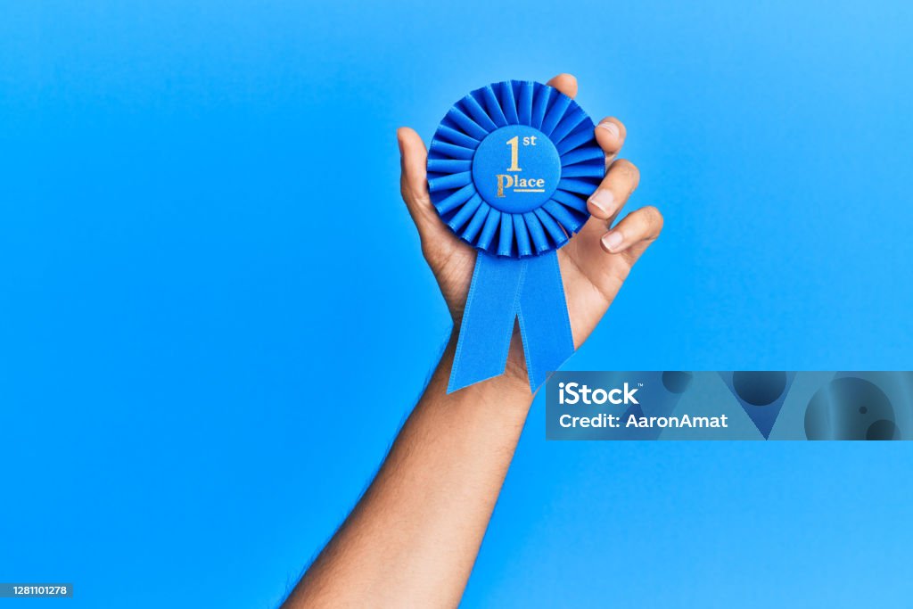 Hand of hispanic man holding 1st place ribbon over isolated blue background. Medal Stock Photo