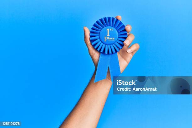 Hand Of Hispanic Man Holding 1st Place Ribbon Over Isolated Blue Background Stock Photo - Download Image Now