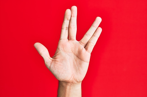 Arm of caucasian white young man over red isolated background greeting doing vulcan salute, showing hand palm and fingers, freak culture