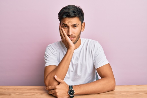 Young handsome man wearing casual white t-shirt sitting on the table thinking looking tired and bored with depression problems with crossed arms.