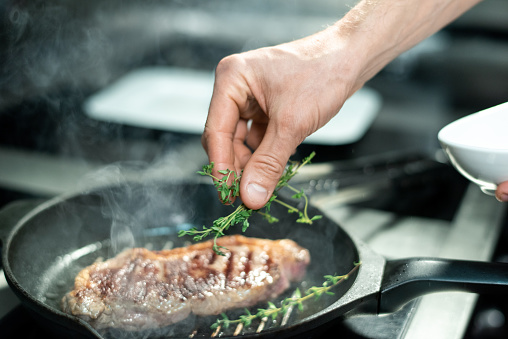 Hand of chef putting green aromatic herbs on roasted piece of meat on hot grill frying pan while standing by electric stove and cooking beef