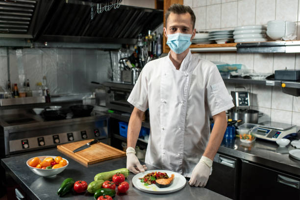 istock Young professional chef in uniform and protective mask holding fried salmon 1281105034