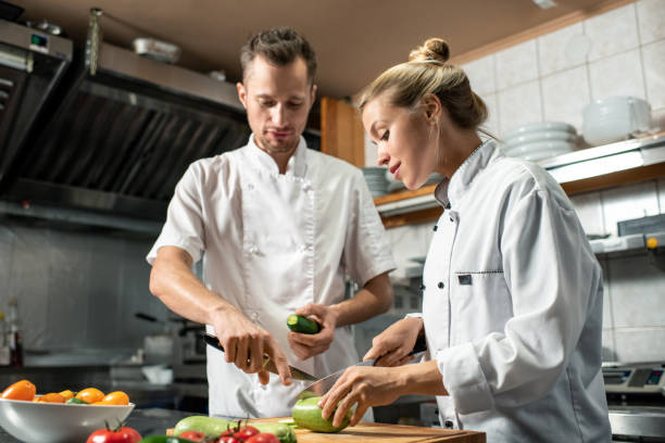 istock Young confident professional chef with knife and cucumber consulting his trainee 1281105035
