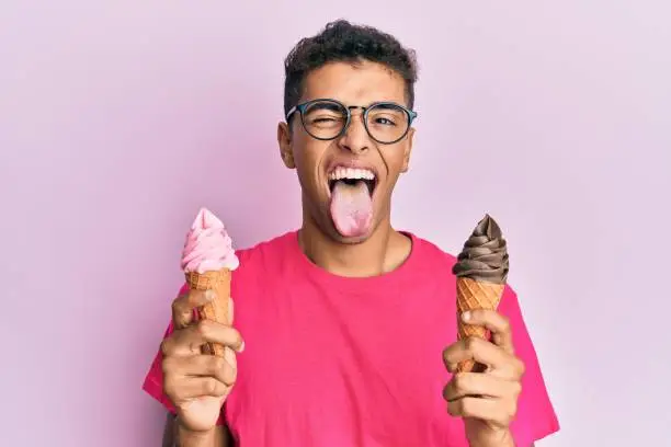 Young handsome african american man holding ice cream cones sticking tongue out happy with funny expression.
