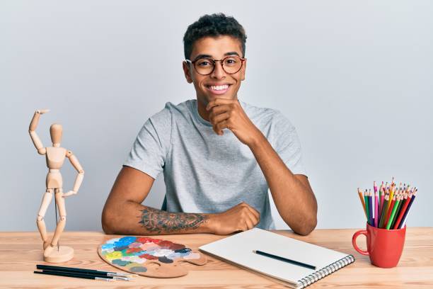 Young handsome african american man painter sitting palette and art manikin looking confident at the camera with smile with crossed arms and hand raised on chin. thinking positive.