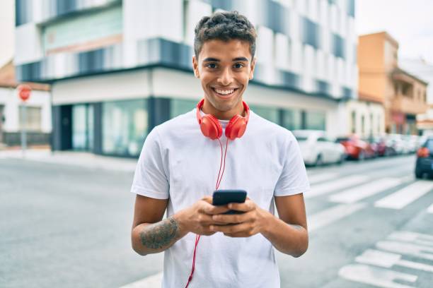 молодой латинский мужчина улыбается счастливым с помощью смартфона и наушников в городе. - city cheerful urban scene happiness стоковые фото и изображения