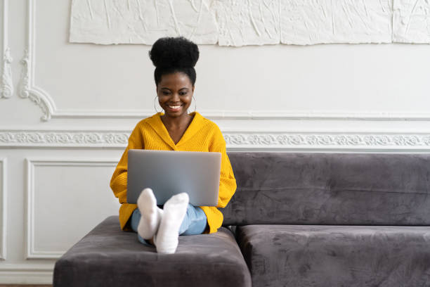 mulher millennial afro-americana sorridente usa cardigan amarelo sentado no sofá, olhando para a webcam da câmera e falando em uma chamada de vídeo ou skype com amigos, assistindo filme - skype - fotografias e filmes do acervo