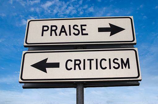 Praise vs criticism. White two street signs with arrow on metal pole with word. Directional road. Crossroads Road Sign, Two Arrow. Blue sky background. Two way road sign with text