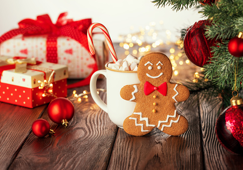 Christmas homemade gingerbread man, candy cane and cup with marshmallow on rustic wooden table. Xmas giftboxes, and yellow defocused lights on background. Defocused fir tree with Christmas balls in front.