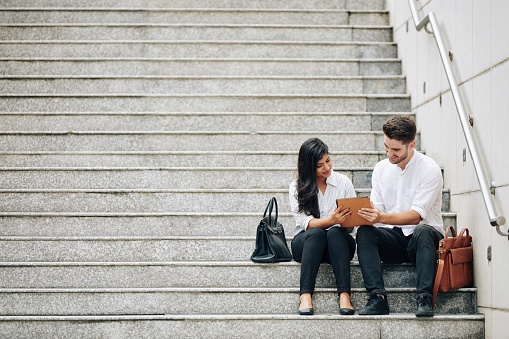 Smiling young multi-ethnic business couple sitting on steps and discussing document on tablet computer