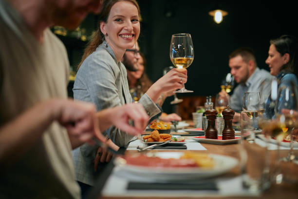 encantadora mujer sonriente sentada en el restaurante con amigos en la cena y sosteniendo una copa de vino blanco. - dining people women wine fotografías e imágenes de stock