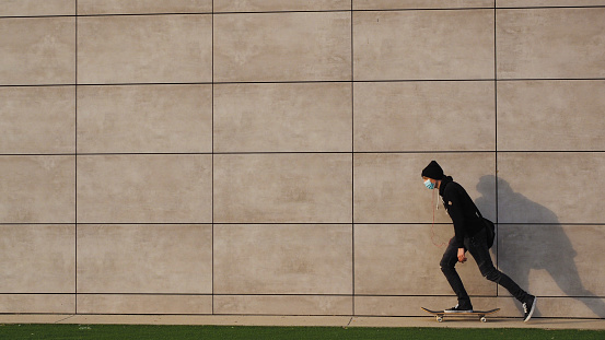 View of a young man skateboarding in the urban environment. The man is casually dressed and wears black jeans and hat. He carries black briefcase hang on his shoulder in front of a gray wall. The shot is executed with available natural light and the copy space has been left. Autumn colors and light.