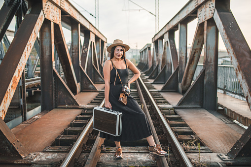 Young beautiful woman tourist in black dress on vacation wearing a hat standing on old rusty bridge carrying a suitcase