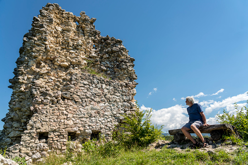 Senior man sitting on the bench watching old stone ruins of castle against blue sky.