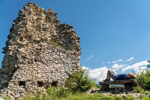 Senior man relaxing on the bench watching old stone ruins of castle against blue sky.